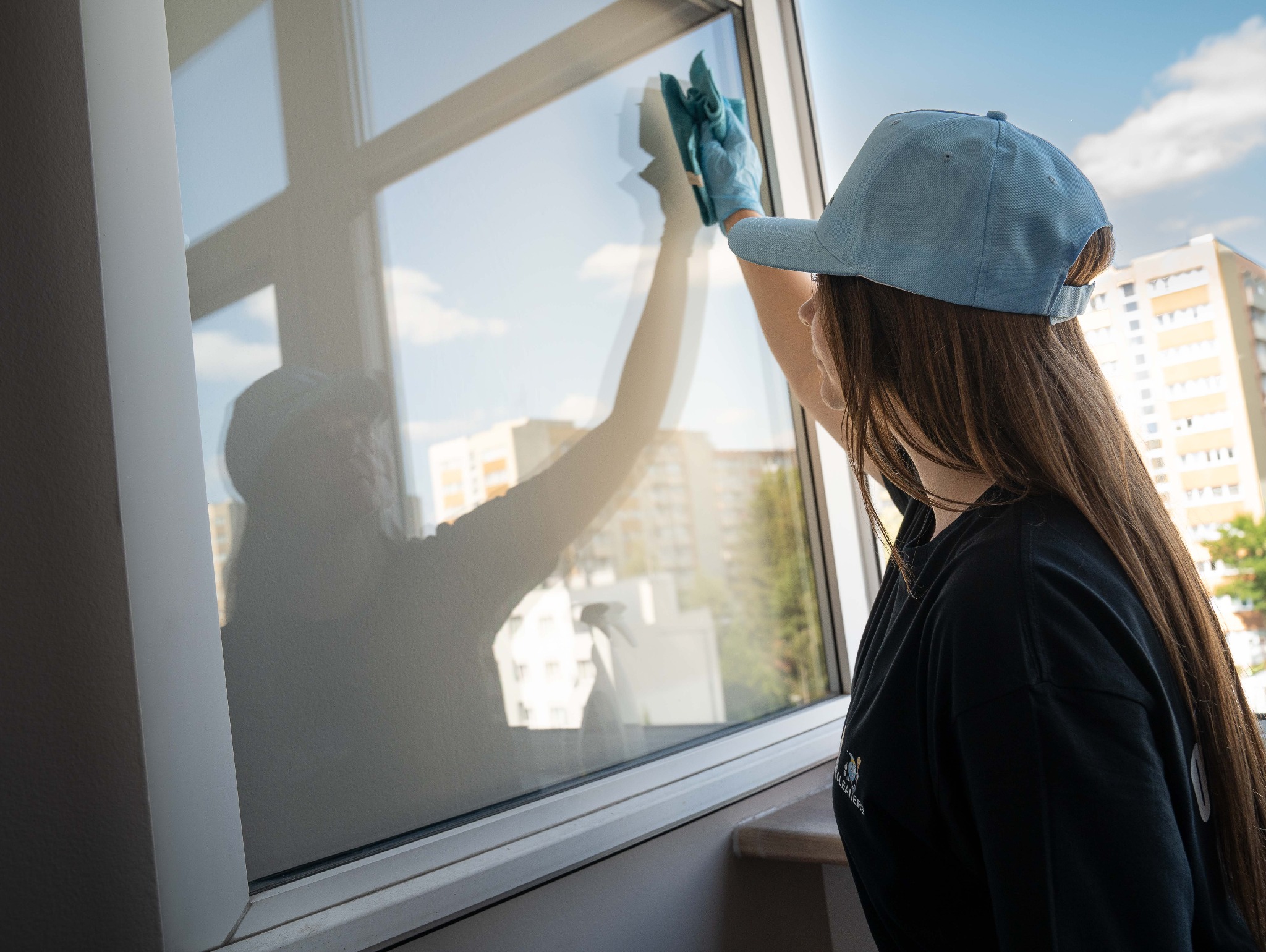 Kobieta w czarnej bluzie i czapce myje okno szmatką w niebieskiej rękawicy. W oknie odbija się sylwetka osoby i budynki na zewnątrz.