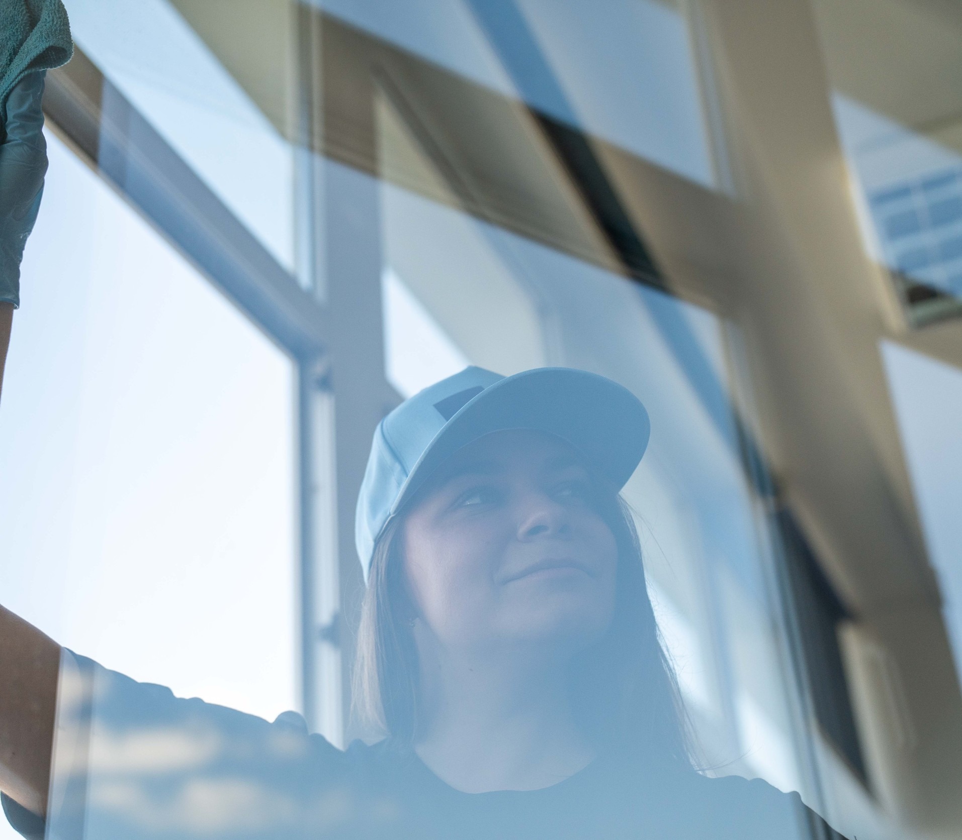 Kobieta w czapce myje okno od wewnątrz, używając szmatki. Widoczne odbicie nieba w czystej szybie, jasne światło dzienne.