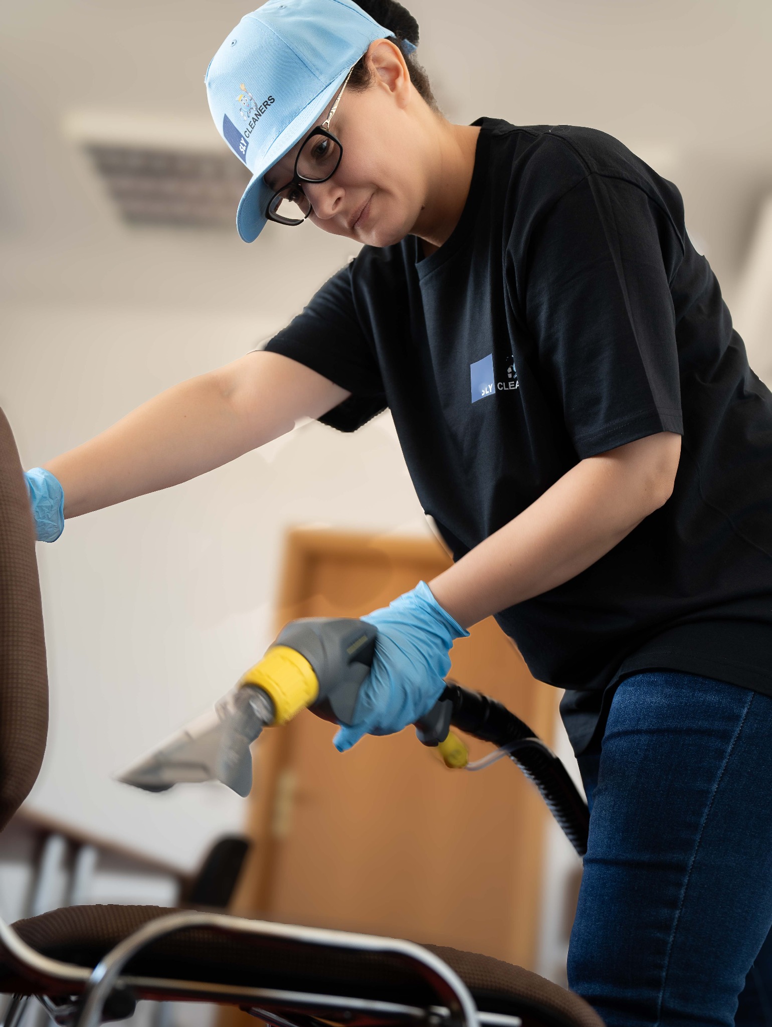 Pracownik w niebieskich rękawicach czyści tapicerkę krzesła biurowego za pomocą profesjonalnego odkurzacza parowego z żółtymi elementami.
