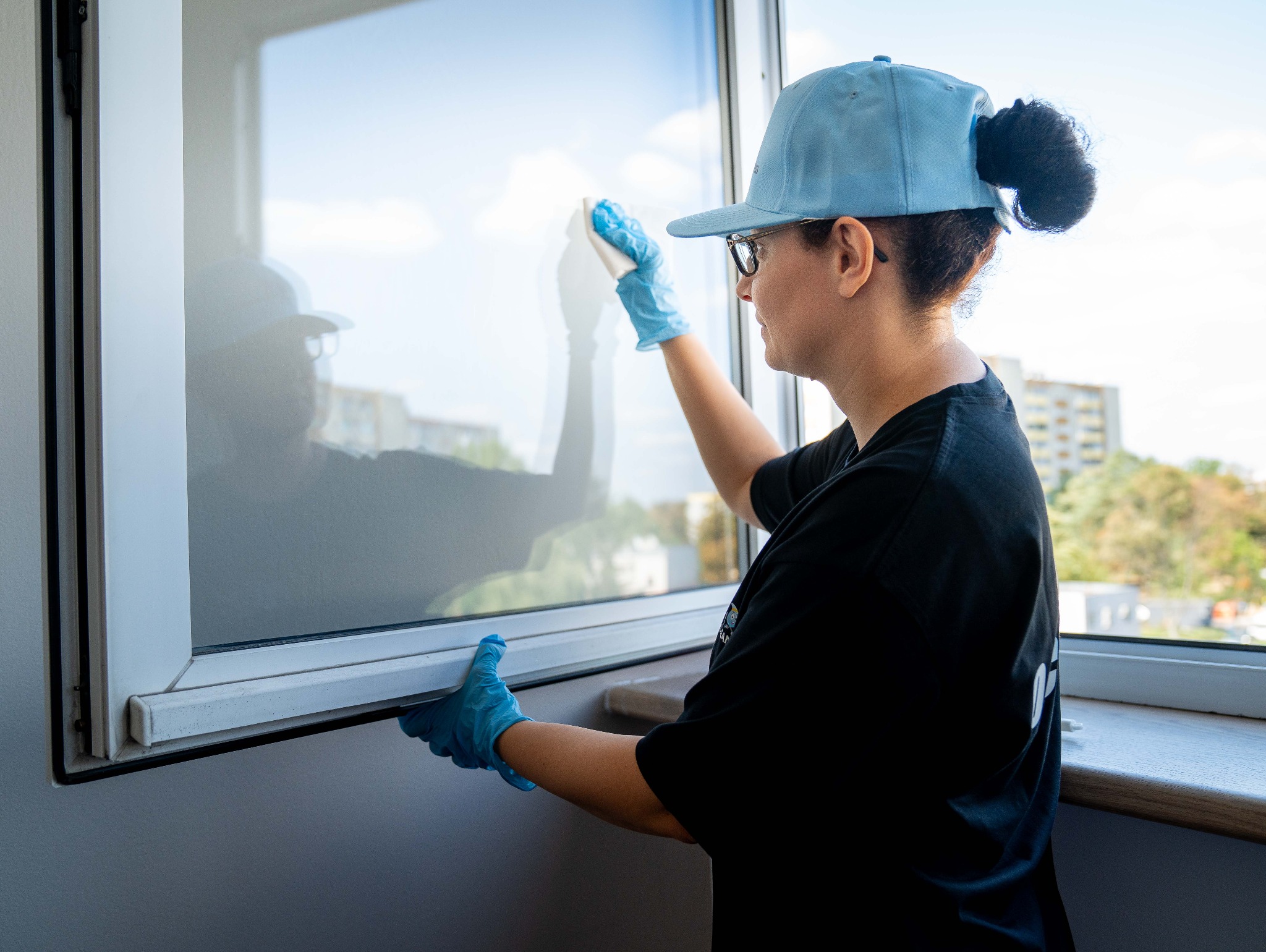 Kobieta w niebieskiej czapce i rękawicach myje okno od wewnątrz, widoczne odbicie w szybie i budynki za oknem.