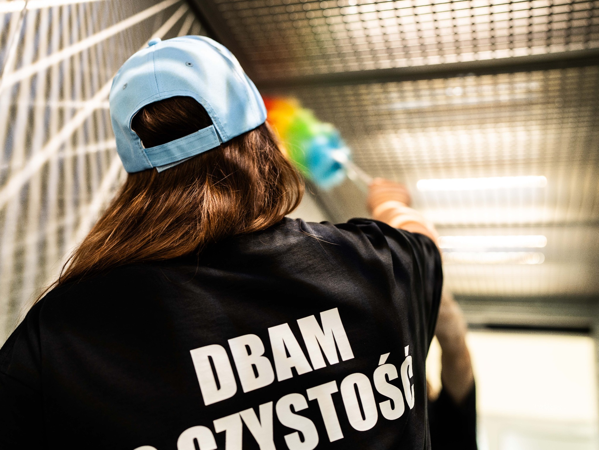 Osoba w czarnej koszulce z napisem 'DBAM O CZYSTOŚĆ' czyści sufit kolorową miotełką. Widoczna błękitna czapka i brązowe włosy. Ujęcie od tyłu, w tle metalowa konstrukcja.