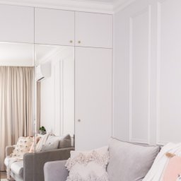 AP Budownictwo Agnieszka Lipińska - Elegancki salon w odcieniach szarości z narożną sofą, ozdobnymi poduszkami i ścianą z dekoracyjnymi listwami. W tle zabudowa szafy z lustrzanymi drzwiami.