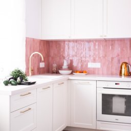 AP Budownictwo Agnieszka Lipińska - Biała kuchnia z różowymi płytkami i złotą armaturą. Uwagę przyciąga parkiet ułożony we wzór jodełki francuskiej oraz świeże zioła na blacie. Jasne, naturalne światło.
