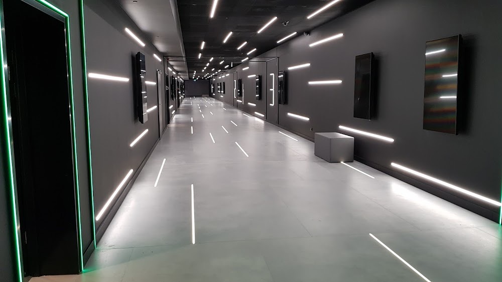 Długi, szary korytarz biurowy z minimalistycznym oświetleniem LED w postaci poziomych pasków na ścianach i suficie. Po prawej stronie widoczne obrazy i siedzisko.