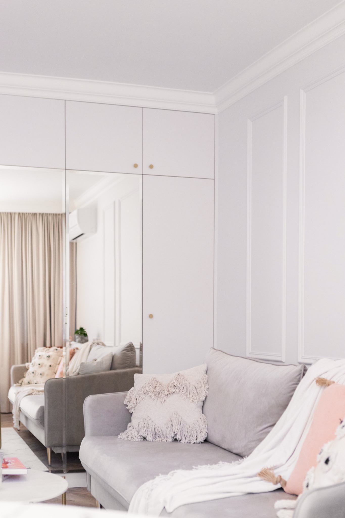 Elegancki salon w odcieniach szarości z narożną sofą, ozdobnymi poduszkami i ścianą z dekoracyjnymi listwami. W tle zabudowa szafy z lustrzanymi drzwiami.