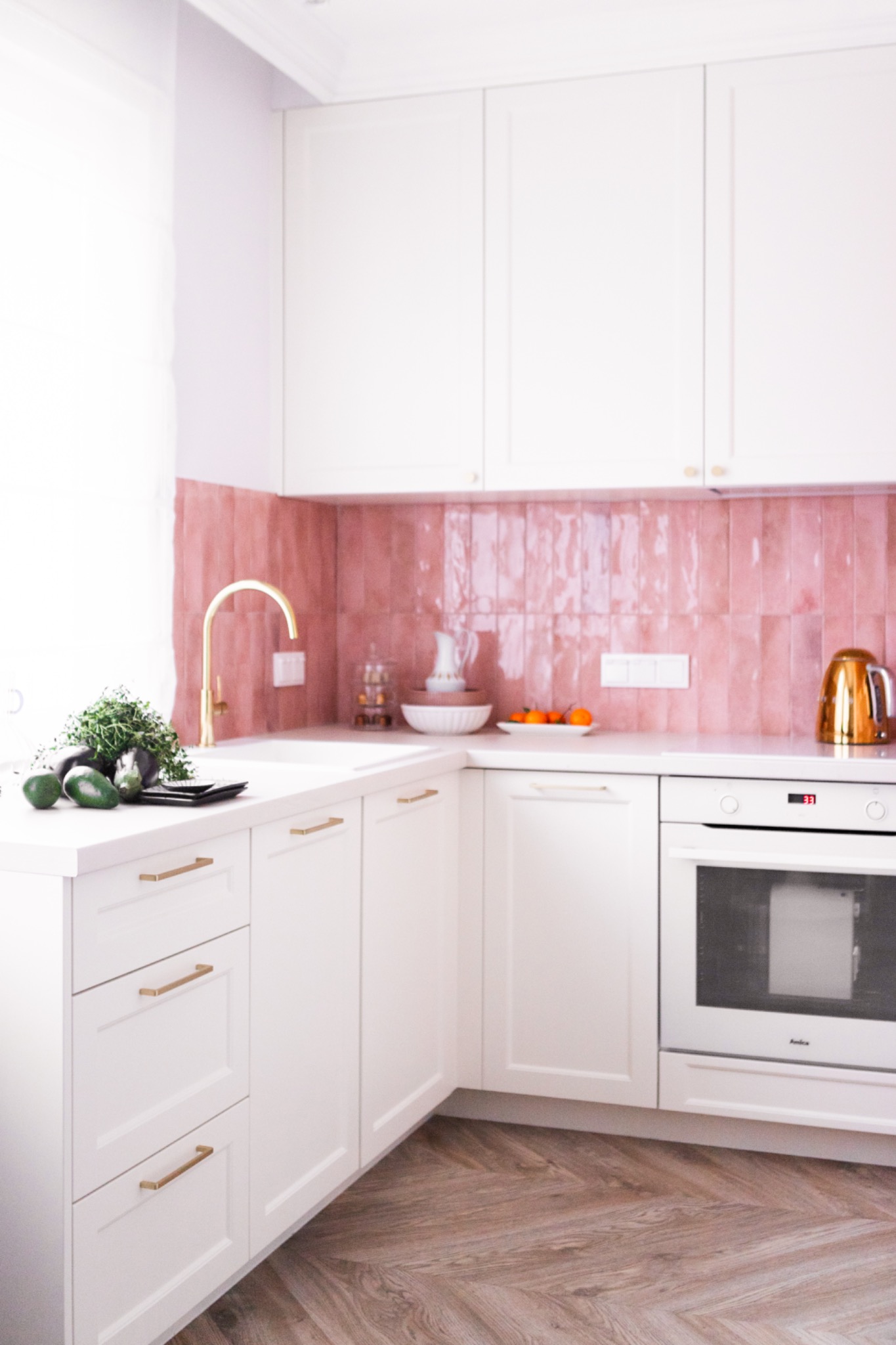 Biała kuchnia z różowymi płytkami i złotą armaturą. Uwagę przyciąga parkiet ułożony we wzór jodełki francuskiej oraz świeże zioła na blacie. Jasne, naturalne światło.