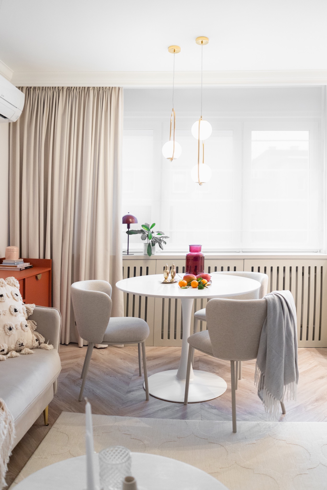 Jasne wnętrze jadalni z okrągłym stołem i minimalistycznymi krzesłami. W tle zasłony i okno z roletami. Na stole owoce i wazon, a nad nim wiszące lampy.