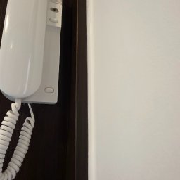 Kamil Dobiesz - Zamontowany domofon w kolorze białym na ciemnym tle drzwi, obok jasnej ściany. Widoczny spiralny kabel. Ujęcie z bliska, skupione na detalu.