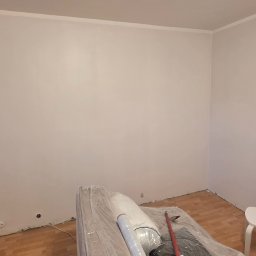 Kamil Dobiesz - Świeżo pomalowane na biało ściany pokoju z widocznymi gniazdkami elektrycznymi, zabezpieczonymi meblami i podłogą. Widoczny biały stołek.