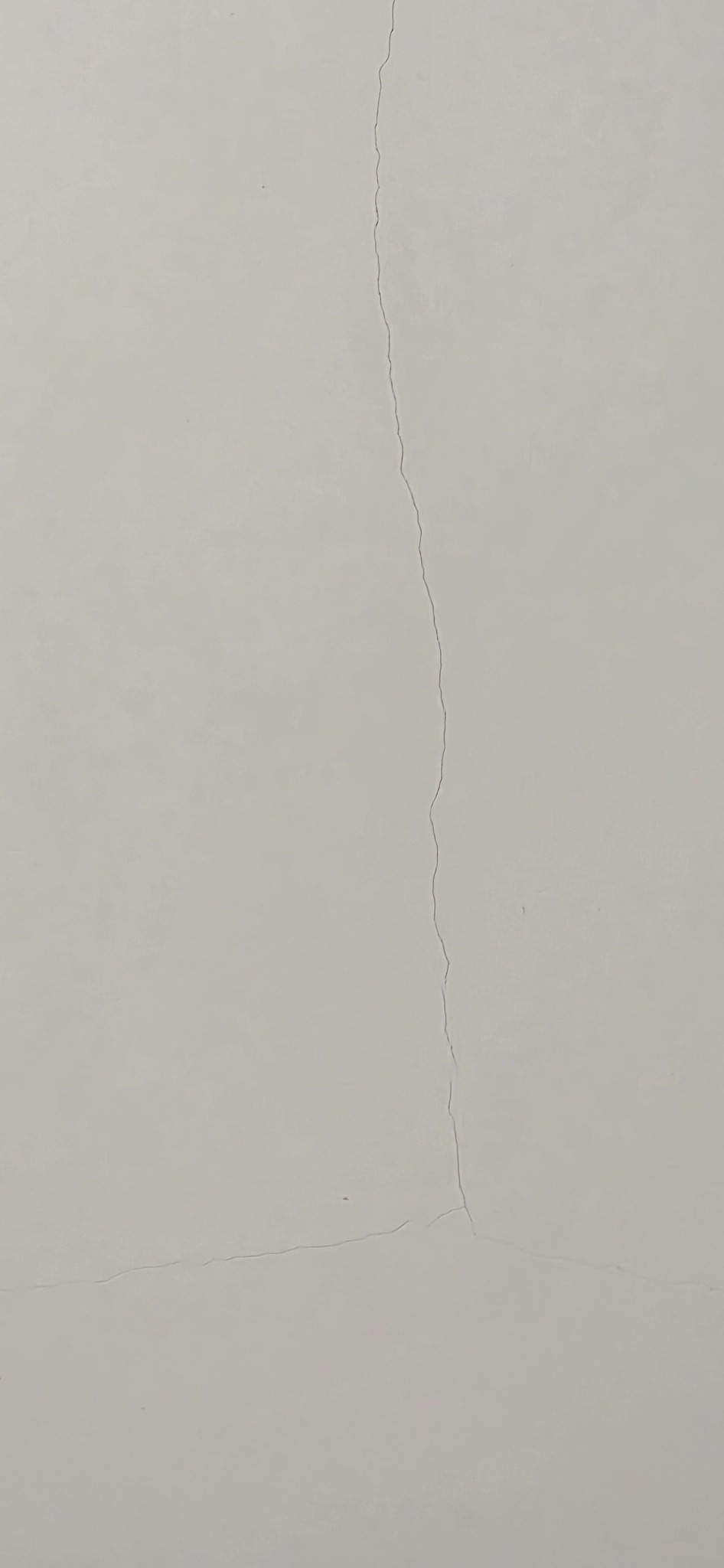 Pęknięcie na gładkiej, jasnej ścianie. Pionowa rysa rozciąga się od góry do dołu, z drobnymi odgałęzieniami w dolnej części. Widoczne delikatne nierówności powierzchni.