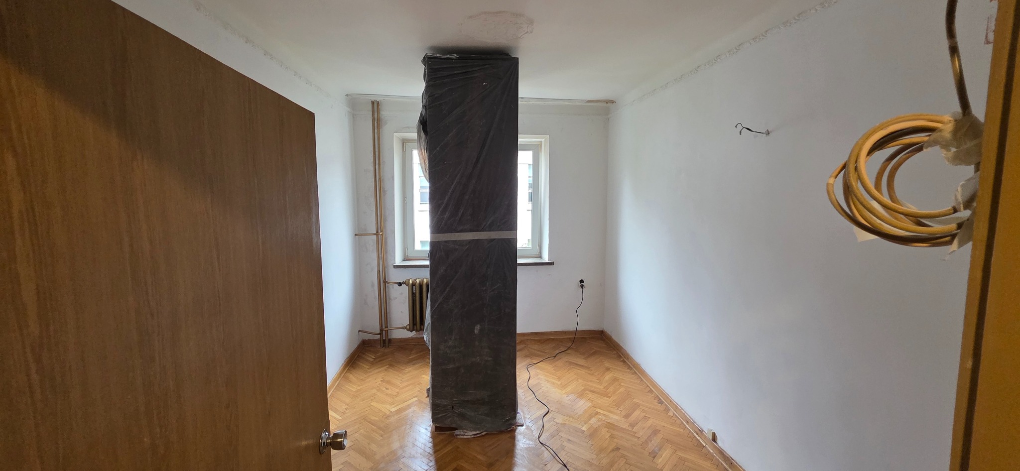 Pokój przed malowaniem: zabezpieczony filar, parkiet, białe ściany z widocznymi śladami po remoncie, grzejnik pod oknem. Przygotowanie do malowania.