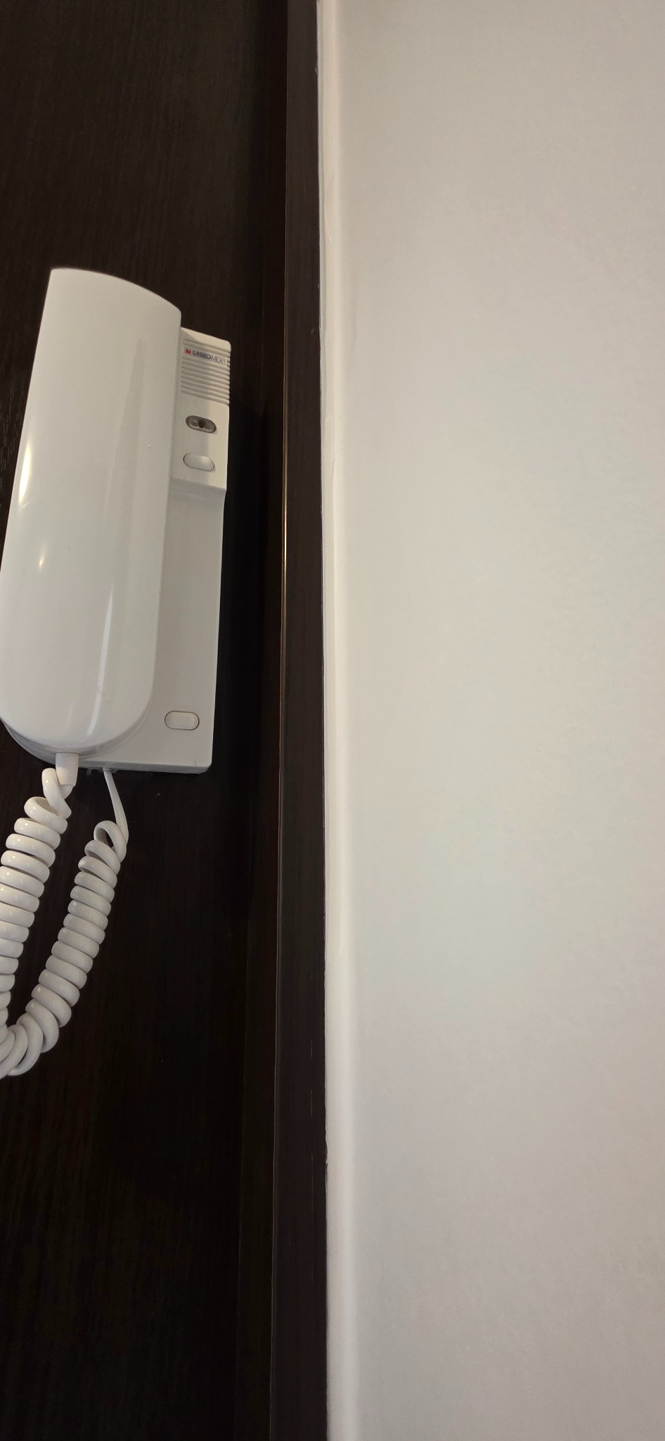 Zamontowany domofon w kolorze białym na ciemnym tle drzwi, obok jasnej ściany. Widoczny spiralny kabel. Ujęcie z bliska, skupione na detalu.