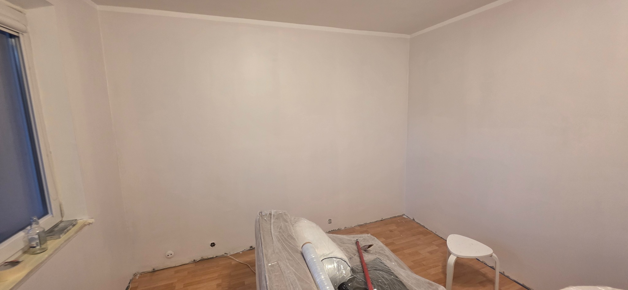 Świeżo pomalowane na biało ściany pokoju z widocznymi gniazdkami elektrycznymi, zabezpieczonymi meblami i podłogą. Widoczny biały stołek.