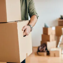 Mężczyzna w oliwkowej koszuli niesie dwa kartony; w tle sterta pudeł, drabina i plecak. Przygotowania do przeprowadzki.