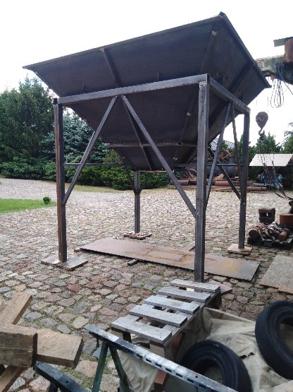 Konstrukcja metalowa z elementami skośnymi, przypominająca zadaszenie, wykonana z surowego metalu, na tle brukowanej powierzchni i zieleni.