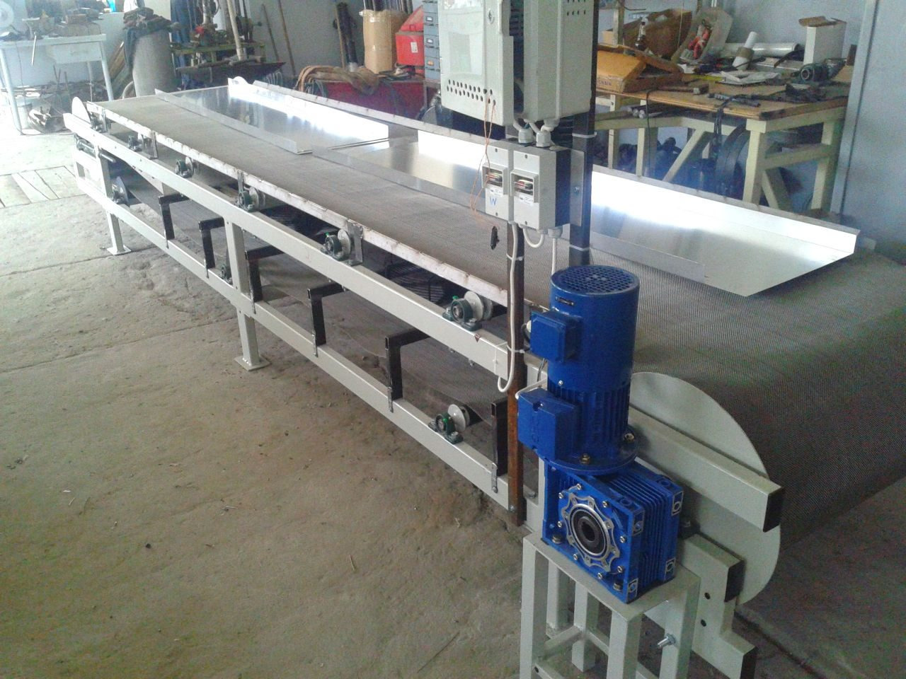 Długa, metalowa konstrukcja z taśmociągiem i niebieskim silnikiem, ustawiona w hali. Widoczne elementy mocujące i podtrzymujące konstrukcję. Szary beton na podłodze.