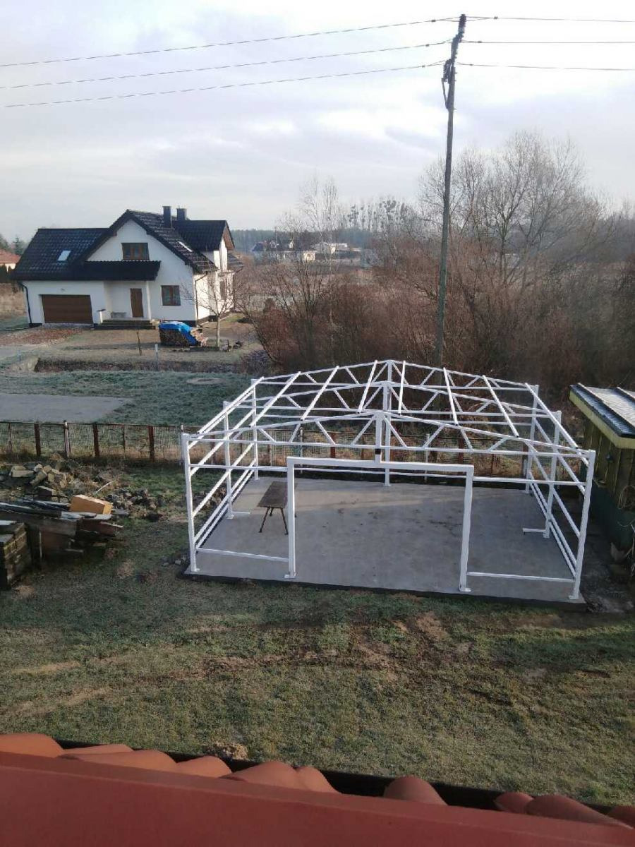 Konstrukcja metalowa pergoli w trakcie montażu, biała rama na betonowej podstawie, widok z góry na tle domu i ogrodu w pochmurny dzień.