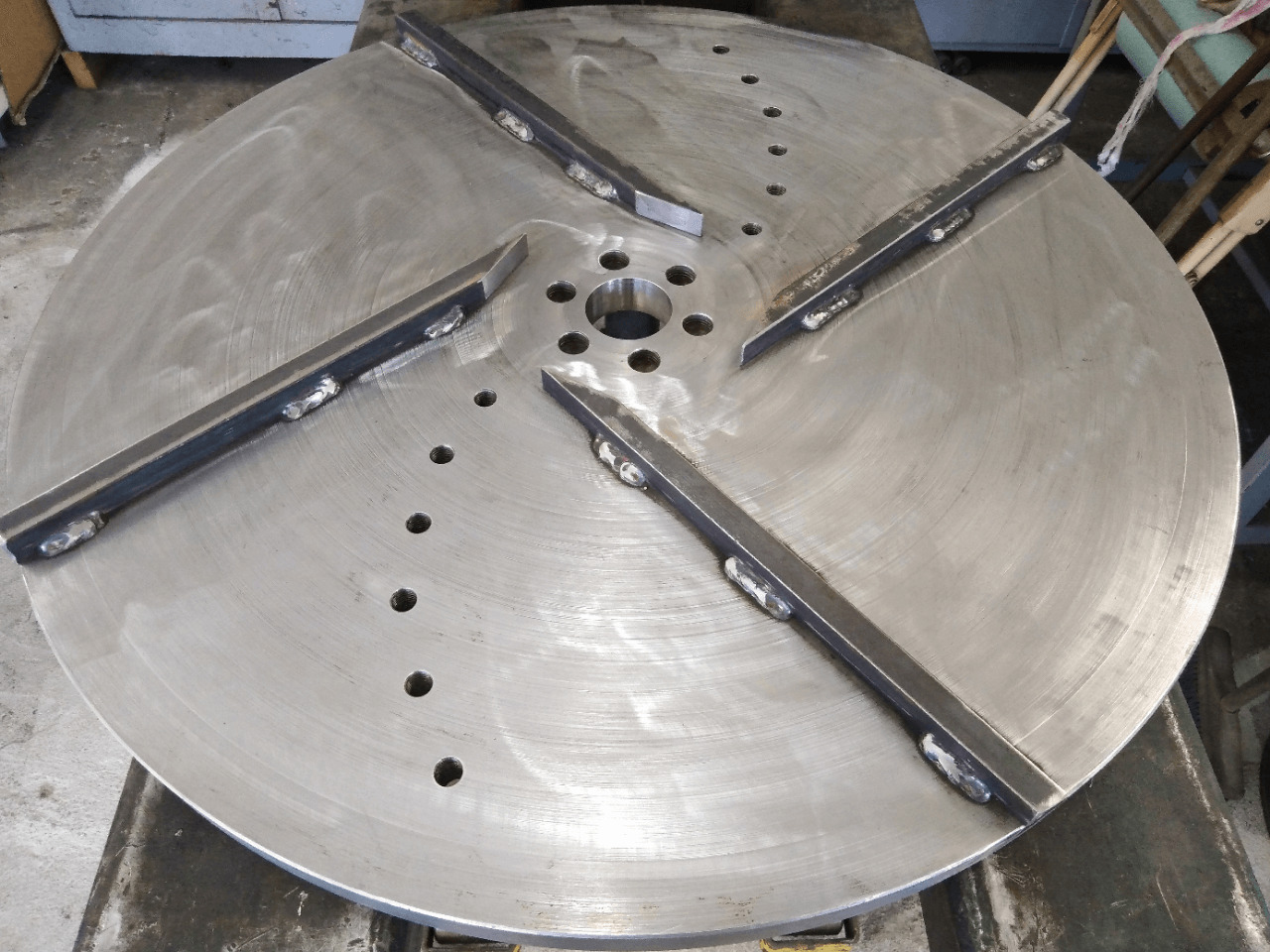 Metalowy dysk z przyspawanymi ostrzami, widoczne ślady obróbki i otwory. Detal konstrukcyjny po spawaniu, surowy metal, przemysłowy charakter.