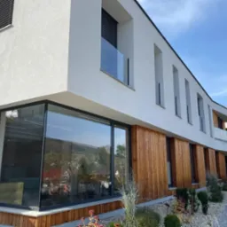 Elewacja nowoczesnego domu z dużymi oknami z czarnymi ramami i drewnianą okładziną. Widok na zielone wzgórza odbijający się w szybach.