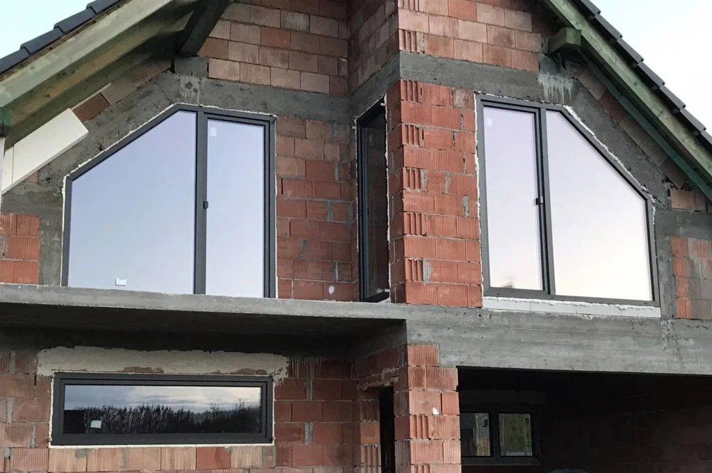 Surowy budynek z czerwonej cegły z zamontowanymi oknami o nietypowych kształtach, w tym trójkątne, w szarych ramach. Widoczne elementy konstrukcyjne.