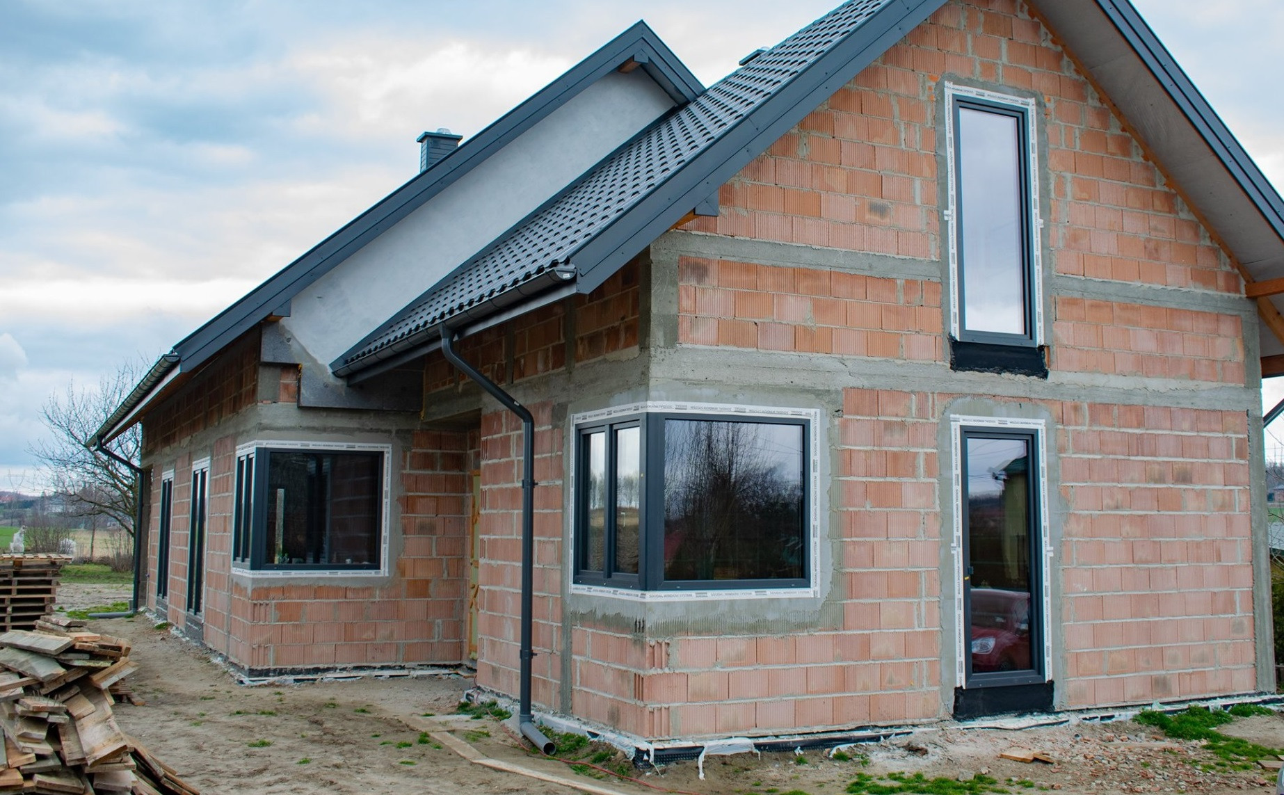 Dom w trakcie budowy z zamontowanymi oknami w ciemnych ramach. Widoczne palety i deski. Dach pokryty szarą dachówką. Częściowo otynkowana elewacja.
