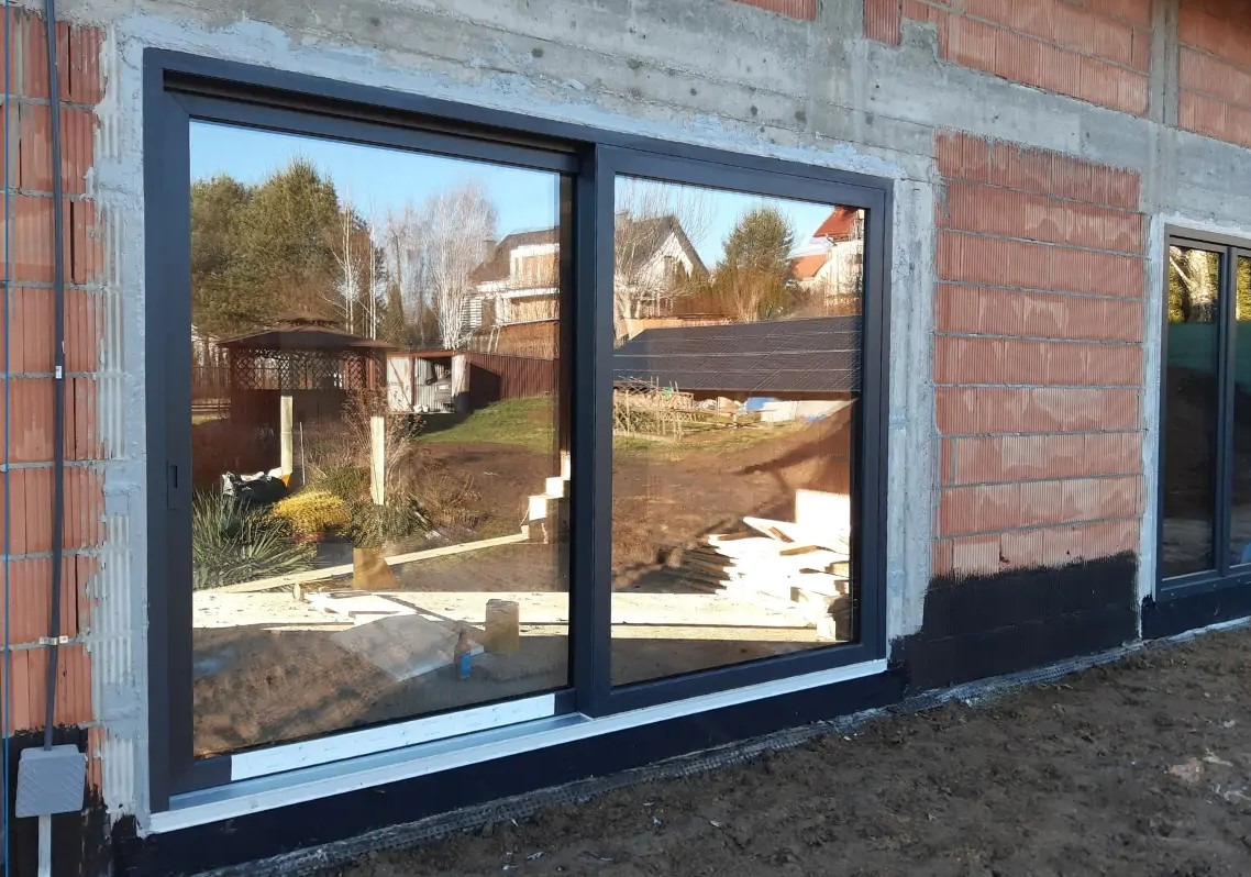 Zamontowane, czarne drzwi przesuwne w surowej ścianie budynku, z widocznym odbiciem ogrodu i domu w szybach. Widoczne detale montażu.