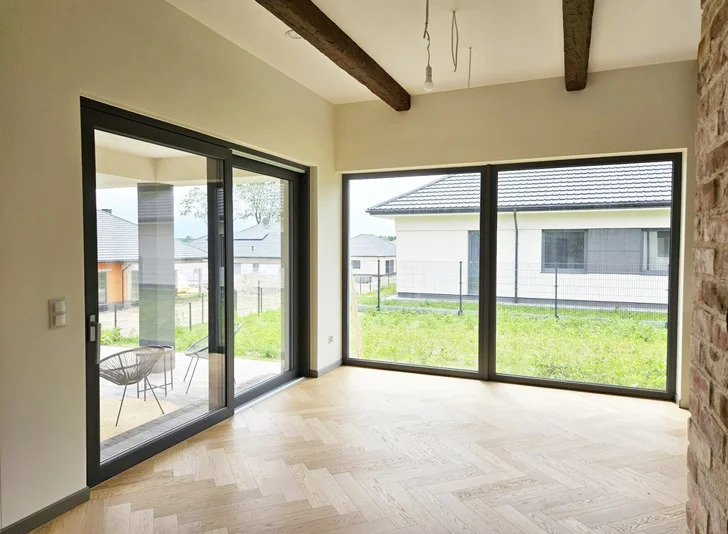 Jasne wnętrze z parkietem w jodełkę, duże okno i przesuwne drzwi balkonowe z widokiem na ogród. W tle domy jednorodzinne.