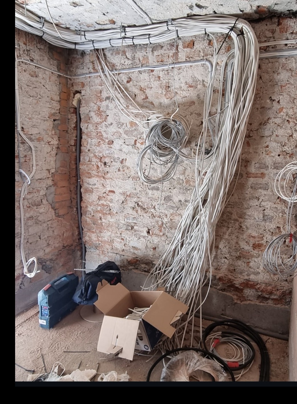 Rozprowadzenie przewodów elektrycznych na ścianie z cegły wewnątrz budynku w trakcie remontu. Widoczne kable, narzędzia i karton. Początek prac domowych.