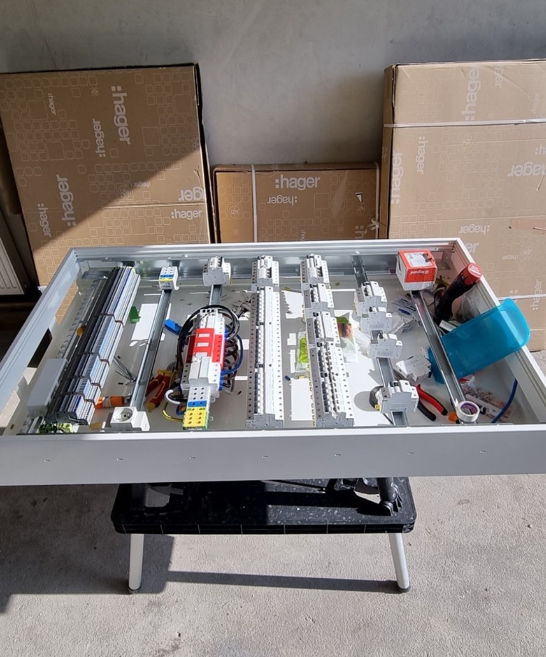 Rozdzielnica elektryczna w trakcie montażu, widoczne elementy instalacji i skrzynki firmy Hager, na tle kartonów. Przewody, bezpieczniki i szyny DIN.