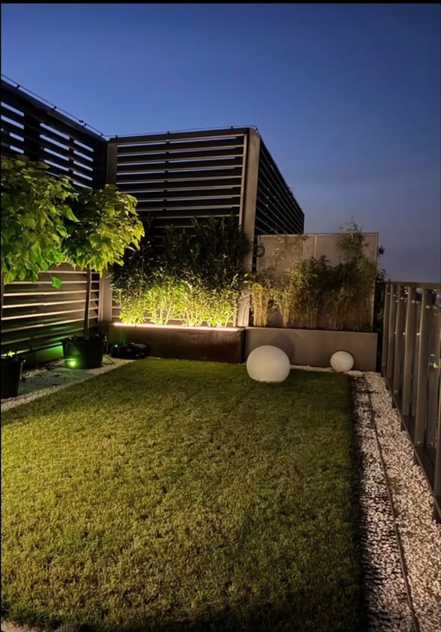 Nowoczesny ogród na dachu z oświetleniem LED pod roślinami, kulami świetlnymi i żwirowym obrzeżem. Widoczne elementy konstrukcji i roślinność.