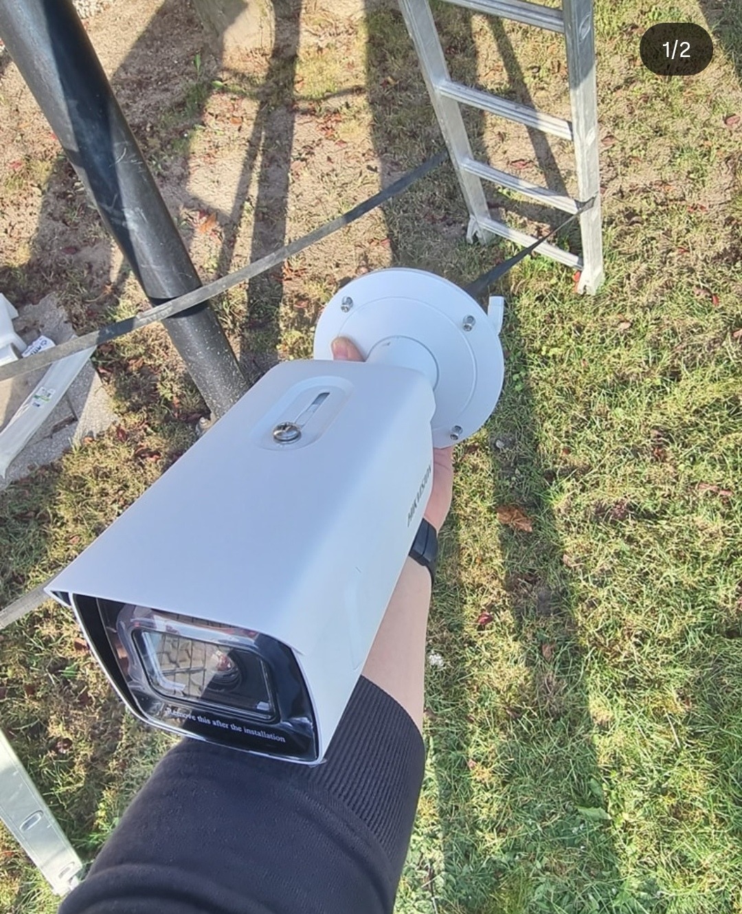 Biała kamera monitoringu Hikvision, trzymana w ręku na tle trawy i drabiny. Widoczny czarny maszt. Przygotowanie do montażu.