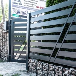GREGOR-STAL mgr inż.Grzegorz Byczkowski - Nowoczesna, pozioma brama wjazdowa z metalowymi przęsłami i gabionowymi słupkami wypełnionymi kamieniem. Widoczny fragment chodnika i zieleni. Tablica informacyjna.