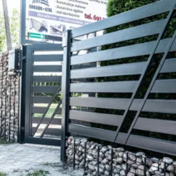 Nowoczesna, pozioma brama wjazdowa z metalowymi przęsłami i gabionowymi słupkami wypełnionymi kamieniem. Widoczny fragment chodnika i zieleni. Tablica informacyjna.