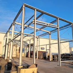 Stalowa konstrukcja hali w trakcie budowy, widoczne słupy i belki, niebieskie niebo w tle, etap montażu szkieletu budynku.