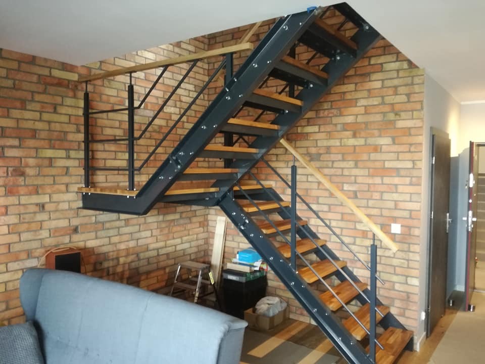 Loftowe schody z metalową konstrukcją i drewnianymi stopniami, umieszczone przy ceglanej ścianie w jasnym wnętrzu. Widoczny fragment sofy.