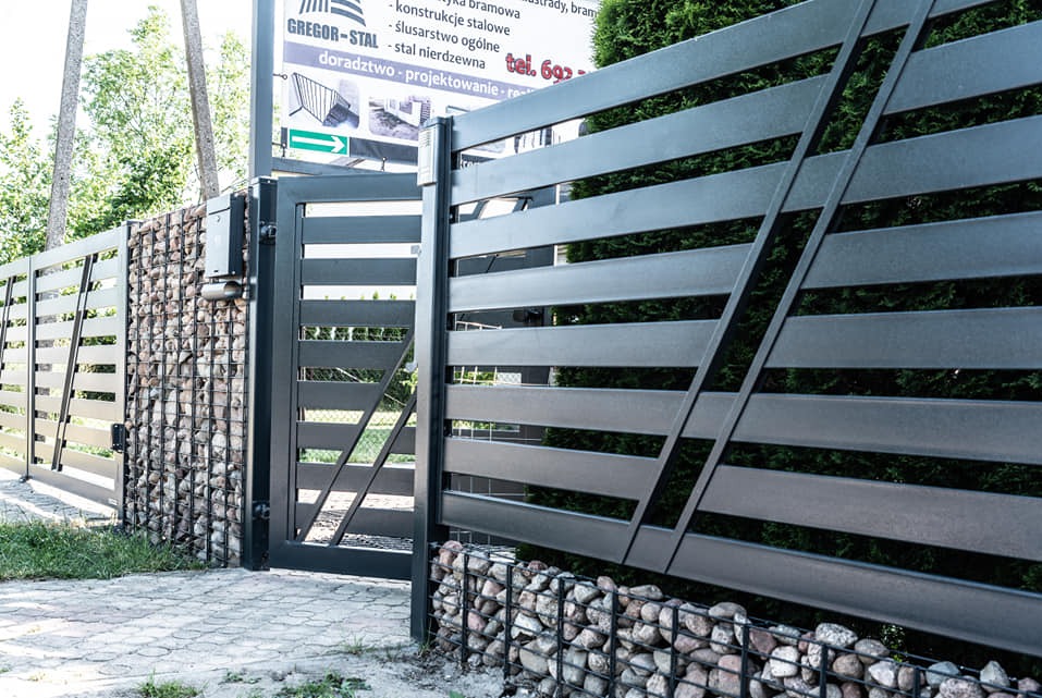 Nowoczesna, pozioma brama wjazdowa z metalowymi przęsłami i gabionowymi słupkami wypełnionymi kamieniem. Widoczny fragment chodnika i zieleni. Tablica informacyjna.