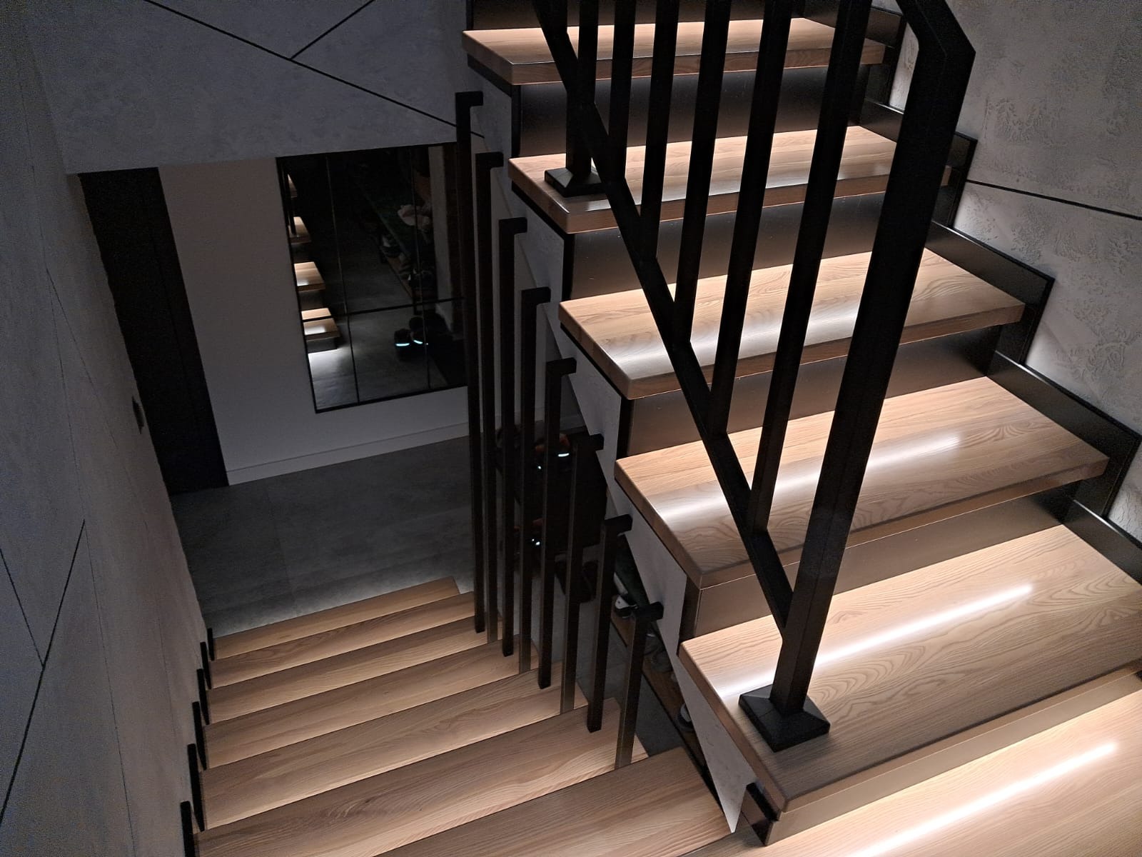 Nowoczesne schody z drewnianymi stopniami podświetlonymi LED i czarną metalową balustradą. Widok z góry, minimalistyczny design, elementy dekoracyjne na ścianie.