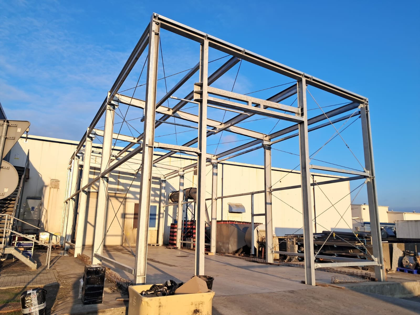 Stalowa konstrukcja hali w trakcie budowy, widoczne słupy i belki, niebieskie niebo w tle, etap montażu szkieletu budynku.