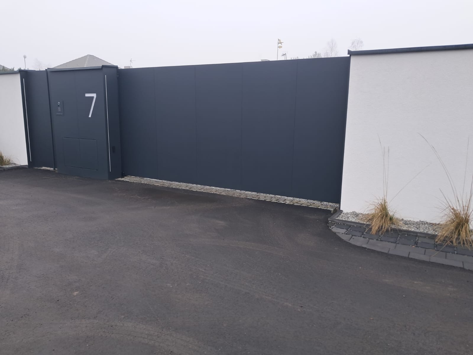 Nowoczesna, przesuwna brama wjazdowa w kolorze antracytowym z numerem posesji '7', osadzona między białymi słupkami. Minimalistyczny design i eleganckie wykończenie.