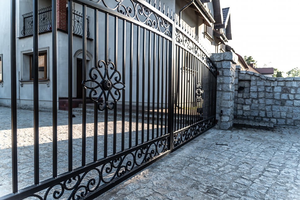 Czarna, kuta brama wjazdowa z dekoracyjnymi rozetami na tle jasnego domu i kamiennego muru. Widoczne cienie i kostka brukowa na podjeździe.