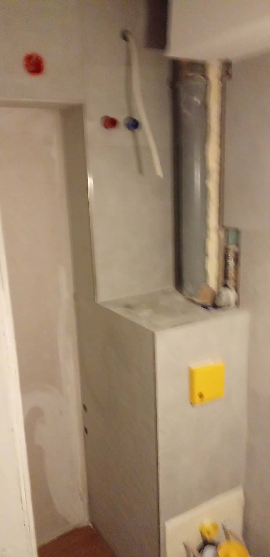 Obudowa rury w łazience wykończona płytkami, widoczne przyłącza wody i kanalizacja. Prace wykończeniowe w trakcie realizacji, pianka montażowa wokół rury.