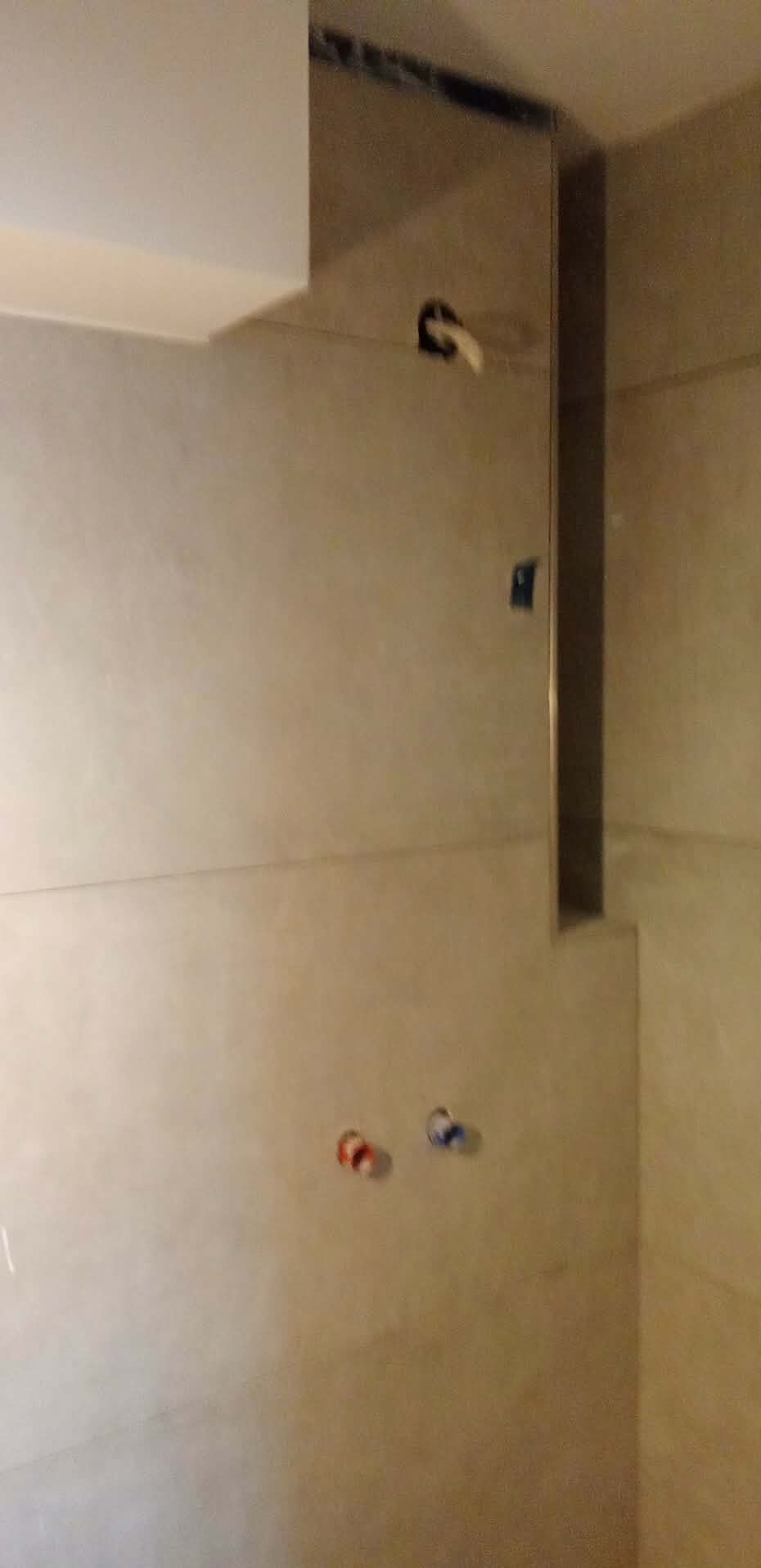 Wykończona ściana w łazience z beżowymi płytkami, widoczne przyłącza wodne i metalowa listwa narożna. Minimalistyczny design, ciepłe oświetlenie.