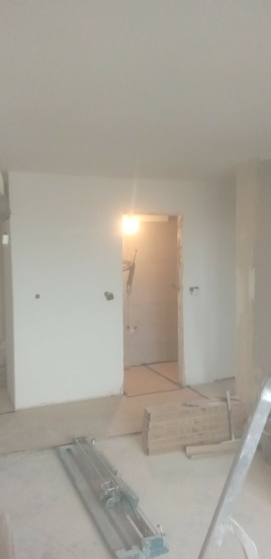 Wnętrze w trakcie remontu: białe ściany, widoczne instalacje, narzędzie do cięcia płytek i ułożone panele podłogowe. Prace wykończeniowe w toku.