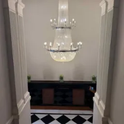 Eleganckie wnętrze z kryształowym żyrandolem, czarno-białą podłogą w romby i kolumnami po bokach. W tle widoczny ciemny bufet z kwiatami.