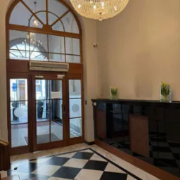 Eleganckie wejście z kryształowym żyrandolem, szachownicą na podłodze i drewnianymi drzwiami z łukowym oknem. Dwie szklane wazy z tulipanami na czarnym blacie.