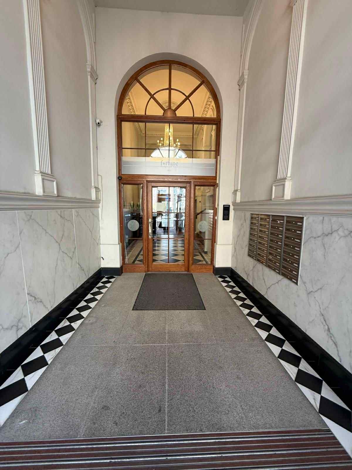Wejście do biura z marmurowymi ścianami, szachownicą na podłodze i skrzynkami pocztowymi. Nad drzwiami widać nazwę firmy 'Fortune'.