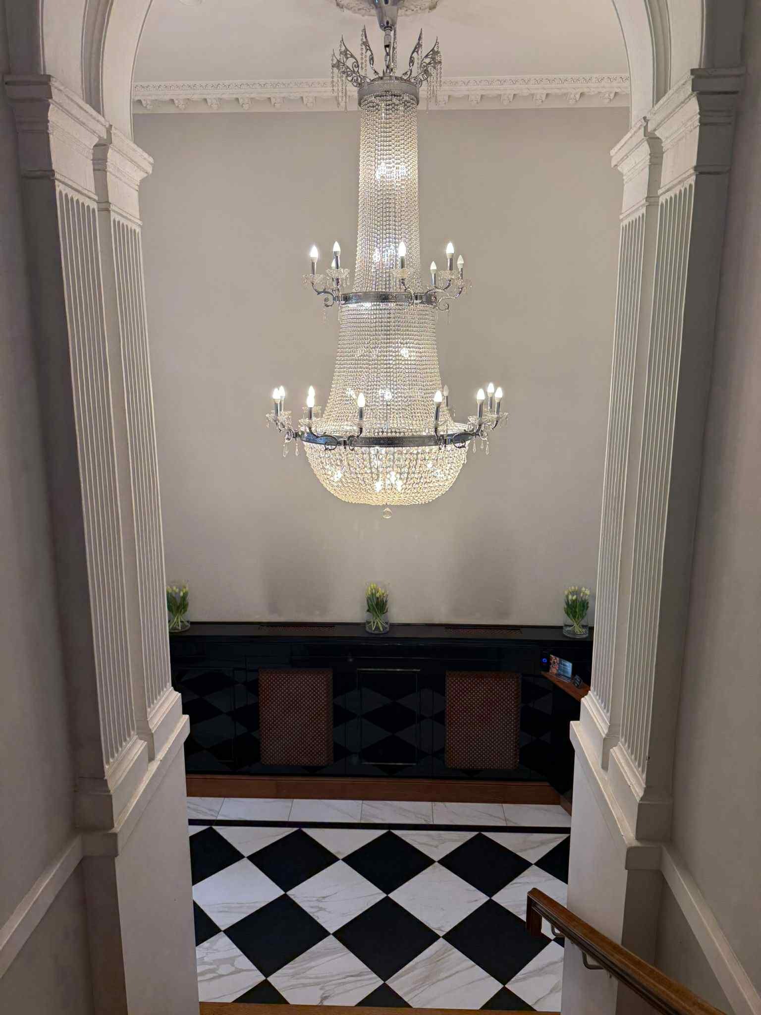 Eleganckie wnętrze z kryształowym żyrandolem, czarno-białą podłogą w romby i kolumnami po bokach. W tle widoczny ciemny bufet z kwiatami.