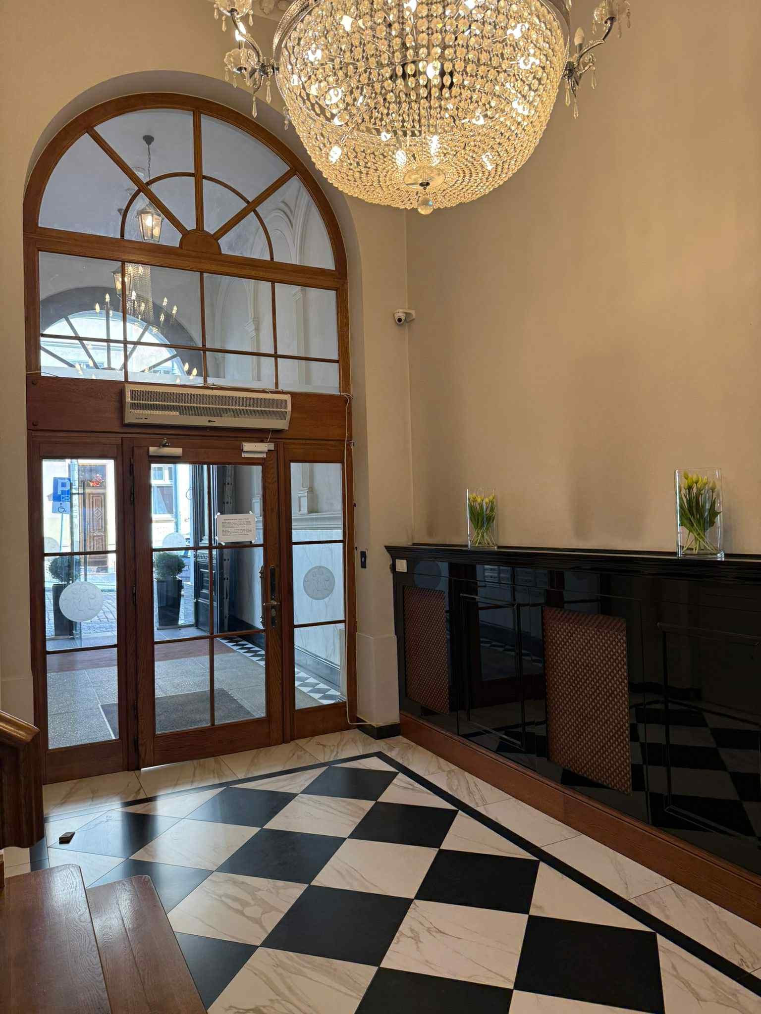 Eleganckie wejście z kryształowym żyrandolem, szachownicą na podłodze i drewnianymi drzwiami z łukowym oknem. Dwie szklane wazy z tulipanami na czarnym blacie.