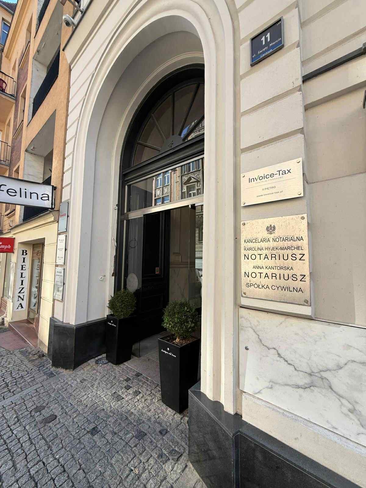Eleganckie wejście do biura Invoice-Tax z szyldem i adresem 'ul. Święty Marcin 11'. Obok wejście do Kancelarii Notarialnej. Kamienny chodnik i rośliny w donicach.
