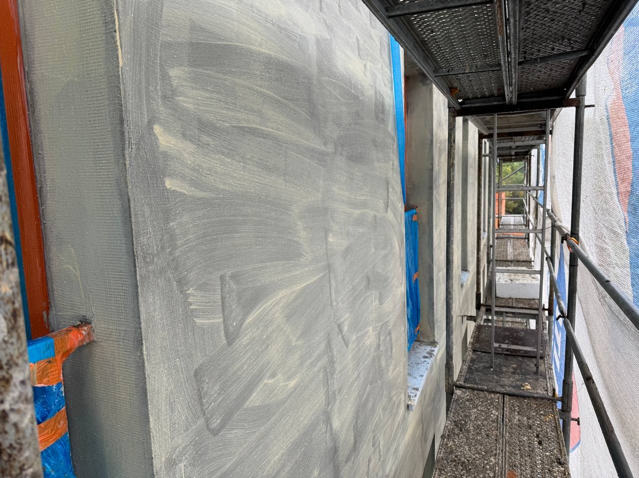 Szara elewacja budynku w trakcie prac wykończeniowych, widoczna siatka i warstwa kleju, rusztowanie z boku. Detal z bliska, podkreślający fakturę i etap prac.